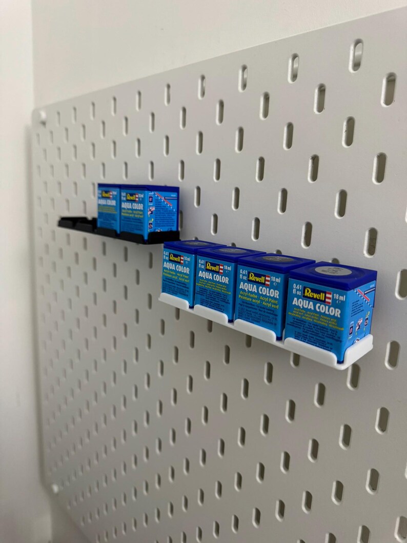 K&ouml;nnte beinhalten: Eine wei&szlig;e Lochwand mit mehreren kleinen blauen Schachteln mit Revell Aqua Color Acrylfarbe. Die Schachteln werden auf schwarzen und wei&szlig;en Regalen ausgestellt. Jede Schachtel enth&auml;lt 18 ml Farbe.