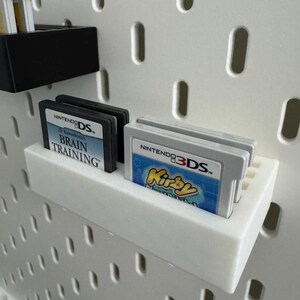 Op de afbeelding: Witte 3D-geprinte planken met Nintendo DS- en 3DS-gamecartridges. E&eacute;n plank toont twee zwarte DS-cartridges met de tekst "Dr. Kawashima's Brain Training". Een andere plank bevat een 3DS-cartridge met de tekst "Kirby".