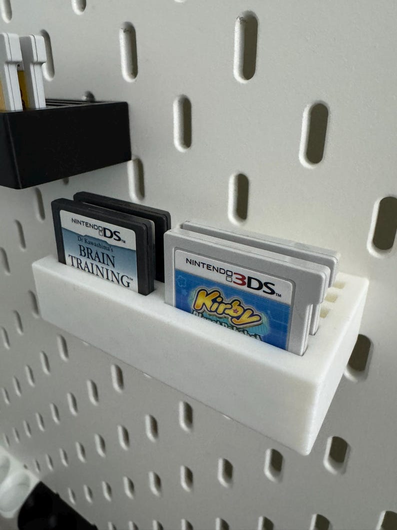 Op de afbeelding: Witte 3D-geprinte planken met Nintendo DS- en 3DS-gamecartridges. E&eacute;n plank toont twee zwarte DS-cartridges met de tekst "Dr. Kawashima's Brain Training". Een andere plank bevat een 3DS-cartridge met de tekst "Kirby".