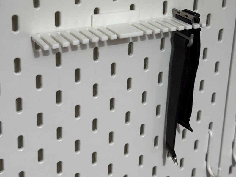 K&ouml;nnte beinhalten: Eine wei&szlig;e Lochwand mit einem schwarzen Gurt, der daran h&auml;ngt. Der Gurt ist an einem wei&szlig;en Kunststoff-Lochwand-Organizer mit mehreren Schlitzen befestigt.