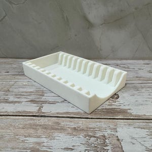 Op de afbeelding: Een witte rechthoekige organizer met een reeks verticale verdelers en kleine vierkante uitsparingen. De organizer is gemaakt van een solide, ondoorzichtig materiaal en staat op een verweerd houten oppervlak.