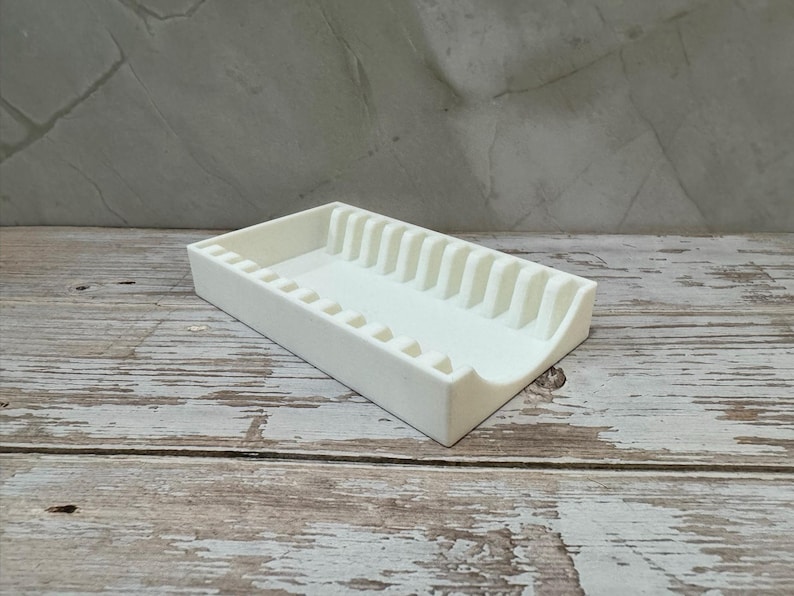 Op de afbeelding: Een witte rechthoekige organizer met een reeks verticale verdelers en kleine vierkante uitsparingen. De organizer is gemaakt van een solide, ondoorzichtig materiaal en staat op een verweerd houten oppervlak.