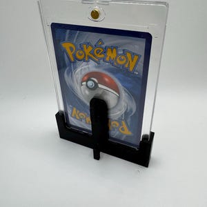 Puede incluir: Un soporte negro que sostiene una carta Pok&eacute;mon dentro de una funda de pl&aacute;stico transparente. La carta presenta el logotipo de Pok&eacute;mon y un dise&ntilde;o de Pok&eacute; Ball rojo y blanco. El soporte se asegura con un cierre dorado.