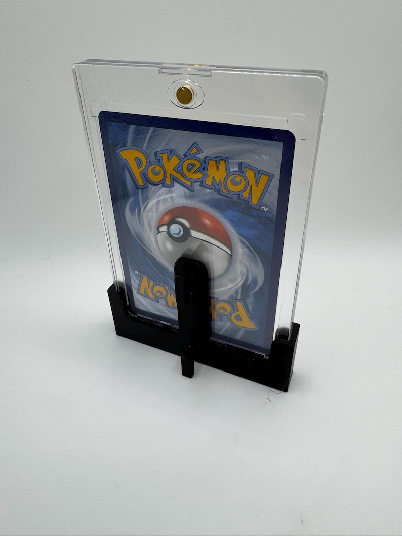 Puede incluir: Un soporte negro que sostiene una carta Pok&eacute;mon dentro de una funda de pl&aacute;stico transparente. La carta presenta el logotipo de Pok&eacute;mon y un dise&ntilde;o de Pok&eacute; Ball rojo y blanco. El soporte se asegura con un cierre dorado.