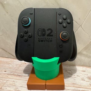 May include: Black Nintendo Switch controller on a green and brown stand. The controller features blue and orange details. The stand is designed to hold the controller upright.