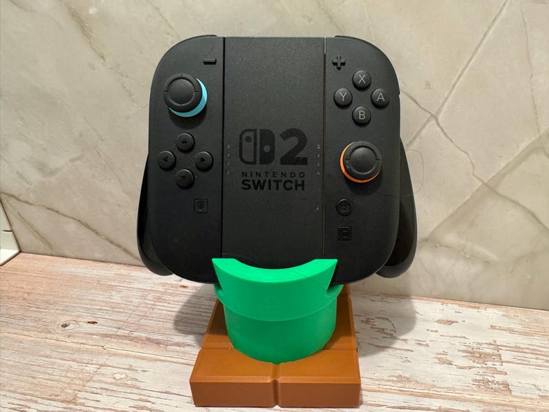 May include: Black Nintendo Switch controller on a green and brown stand. The controller features blue and orange details. The stand is designed to hold the controller upright.
