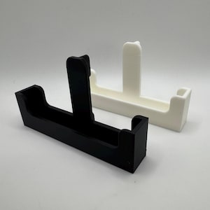 May include: Two minimalist, geometric phone stands. One is black, the other is white. Each stand has a rectangular base with a vertical support and a curved top. The stands are made of a smooth, solid material.