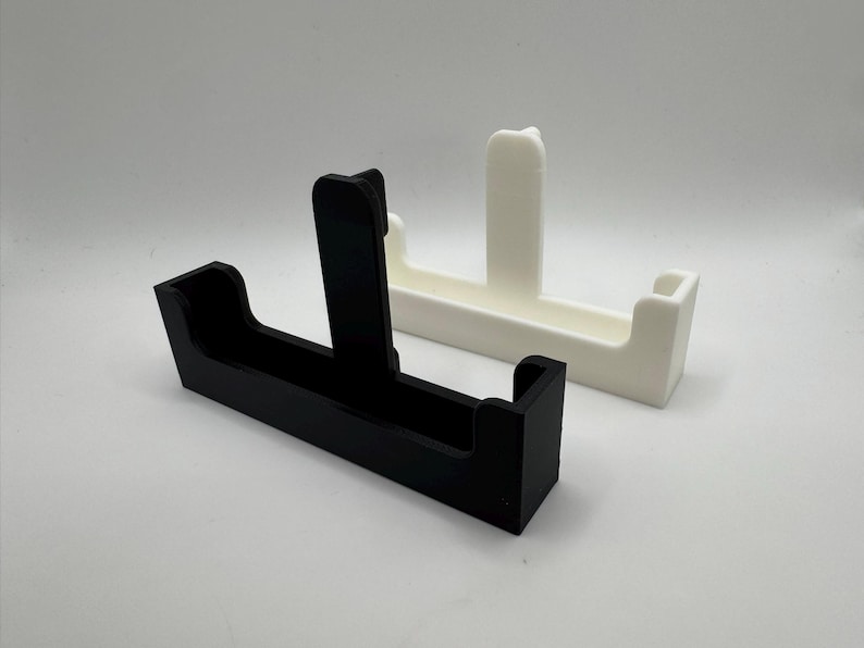 May include: Two minimalist, geometric phone stands. One is black, the other is white. Each stand has a rectangular base with a vertical support and a curved top. The stands are made of a smooth, solid material.