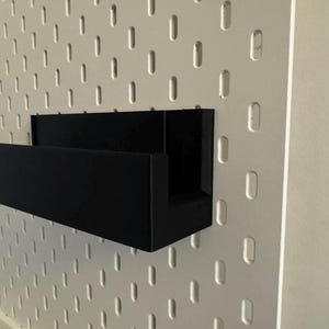 Peut inclure: &Eacute;tag&egrave;re noire rectangulaire fix&eacute;e &agrave; un mur en panneau perfor&eacute; blanc. L'&eacute;tag&egrave;re a un profil en forme de U. Le panneau perfor&eacute; pr&eacute;sente des trous ovales espac&eacute;s. Le mur est de couleur neutre.