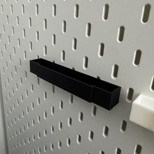 K&ouml;nnte beinhalten: Eine schwarze, rechteckige Aufbewahrungsbox an einer wei&szlig;en Lochwand. Die Box ist f&uuml;r kleine Gegenst&auml;nde konzipiert. Die Lochwand hat gleichm&auml;&szlig;ig verteilte ovale L&ouml;cher.