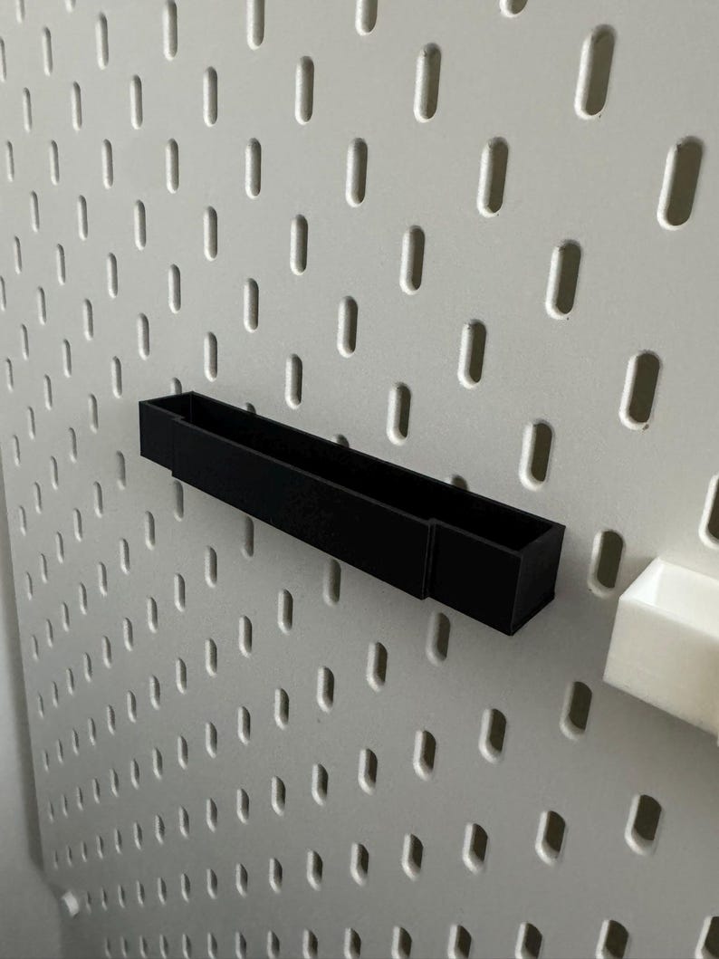 K&ouml;nnte beinhalten: Eine schwarze, rechteckige Aufbewahrungsbox an einer wei&szlig;en Lochwand. Die Box ist f&uuml;r kleine Gegenst&auml;nde konzipiert. Die Lochwand hat gleichm&auml;&szlig;ig verteilte ovale L&ouml;cher.