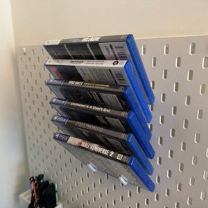 May include: A stack of six PS4 video game cases with blue spines, displayed on a white pegboard. The game titles include "BLADE", "Call of Duty: Infinite Warfare", "Uncharted 4: A Thief's End", "Dishonored 2", and "Dragon Ball Xenoverse 2".