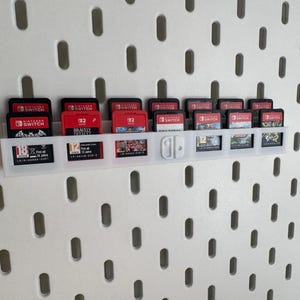 K&ouml;nnte beinhalten: Ein wei&szlig;er Spielkassettenhalter aus Kunststoff, der an einer wei&szlig;en Lochwand befestigt ist. Der Halter enth&auml;lt mehrere rote und schwarze Nintendo Switch-Spielkassetten. Einige Kassetten sind mit Spieletiteln und Nummern beschriftet.