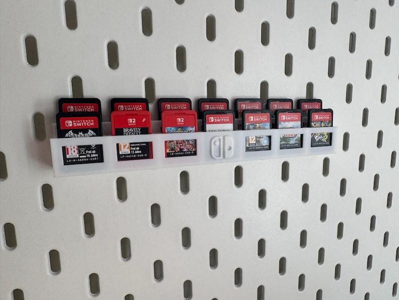 K&ouml;nnte beinhalten: Ein wei&szlig;er Spielkassettenhalter aus Kunststoff, der an einer wei&szlig;en Lochwand befestigt ist. Der Halter enth&auml;lt mehrere rote und schwarze Nintendo Switch-Spielkassetten. Einige Kassetten sind mit Spieletiteln und Nummern beschriftet.