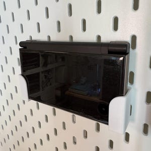 May include: A black handheld gaming console is mounted on a white pegboard using a white bracket. The console is in the closed position, with a reflective screen. The pegboard has evenly spaced oval holes.