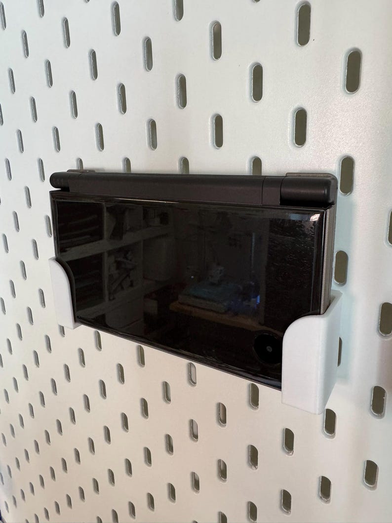 May include: A black handheld gaming console is mounted on a white pegboard using a white bracket. The console is in the closed position, with a reflective screen. The pegboard has evenly spaced oval holes.