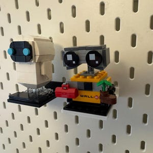 May include: A white and grey robot with blue eyes and a black base, and a yellow and grey robot with black eyes and a black base, both robots are holding objects, the yellow robot is holding a plant and a red toolbox, the white robot is holding a black object, both robots are on a white pegboard.