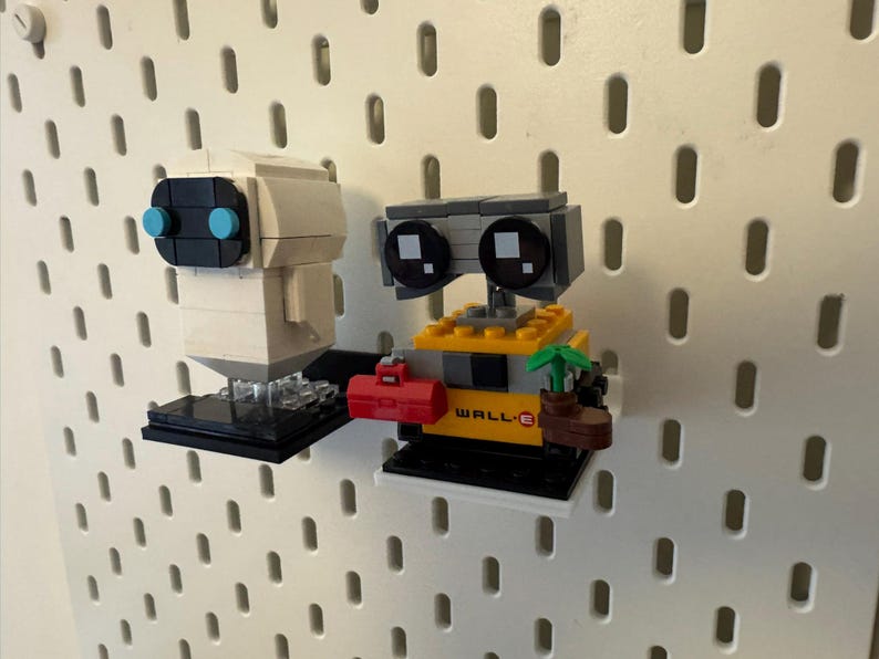 May include: A white and gray robot with blue eyes and a black base, and a yellow and gray robot with black eyes and a black base, both robots are holding objects, the yellow robot is holding a plant and a red toolbox, the white robot is holding a black object, both robots are on a white pegboard.