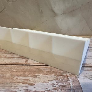 Peut inclure: Un objet rectangulaire en bois de couleur claire avec un dessus inclin&eacute;. L'objet est positionn&eacute; en biais sur une surface en bois. L'arri&egrave;re-plan pr&eacute;sente un mur textur&eacute; de couleur claire.