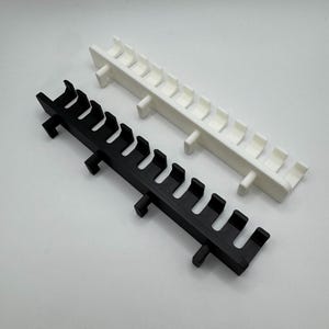 May include: Two plastic, rack-like objects. One is black, the other white. Each has a series of curved, hook-like protrusions along its length, and small rectangular supports. The objects are set against a plain, light background.