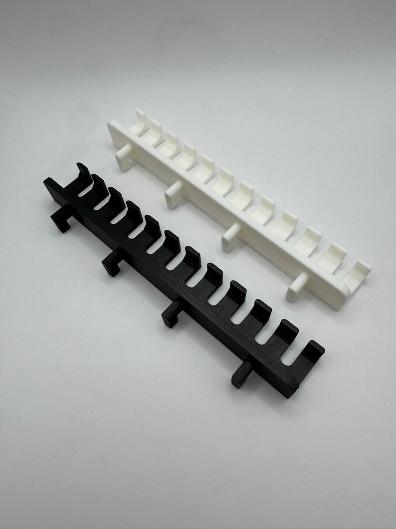 May include: Two plastic, rack-like objects. One is black, the other white. Each has a series of curved, hook-like protrusions along its length, and small rectangular supports. The objects are set against a plain, light background.