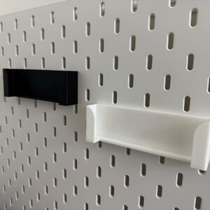 Pu&ograve; includere: Due mensole rettangolari, una nera e una bianca, sono montate su una pannello forato bianco. Le mensole hanno un design semplice e minimalista e sono realizzate in un materiale solido. Il pannello forato ha fori distanziati uniformemente.