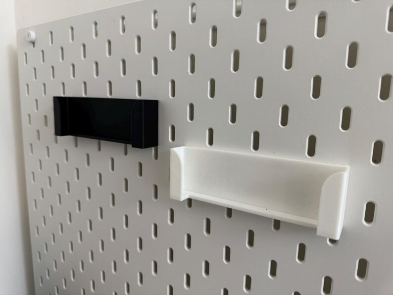 Pu&ograve; includere: Due mensole rettangolari, una nera e una bianca, sono montate su una pannello forato bianco. Le mensole hanno un design semplice e minimalista e sono realizzate in un materiale solido. Il pannello forato ha fori distanziati uniformemente.