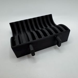 以下が含まれることがあります： 黒色の3Dプリントオーガナイザー。上部は湾曲しており、複数の垂直スロットがあります。前面からは2つの円筒形のペグが伸びています。このオーガナイザーは、机やワークスペースでアイテムを保持し、表示するように設計されています。