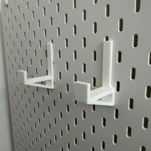 May include: Two white pegboard hooks are mounted on a white pegboard. The hooks are L-shaped and designed to hold items. The pegboard has a grid of evenly spaced holes.