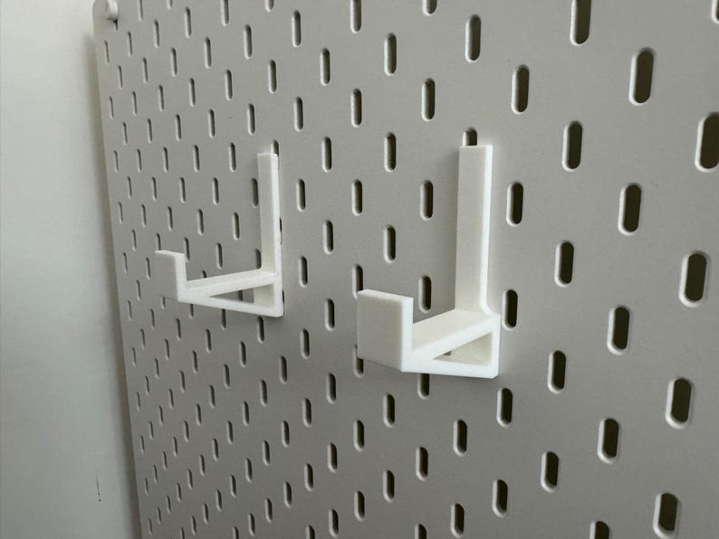 May include: Two white pegboard hooks are mounted on a white pegboard. The hooks are L-shaped and designed to hold items. The pegboard has a grid of evenly spaced holes.