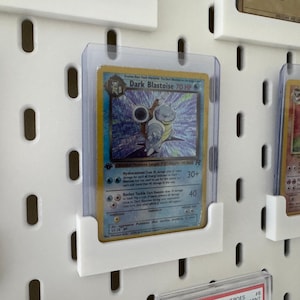 Pu&ograve; includere: Un pannello forato bianco con diverse carte Pokemon esposte in buste di plastica trasparente. Le carte sono tenute in posizione da supporti in plastica bianca. Le carte includono Dark Blastoise, Flareon e una terza carta con il testo "2000 Pokemon Misty's Sea King Holo Rare".