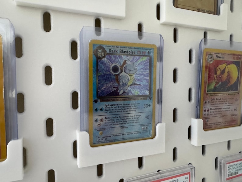 Pu&ograve; includere: Un pannello forato bianco con diverse carte Pokemon esposte in buste di plastica trasparente. Le carte sono tenute in posizione da supporti in plastica bianca. Le carte includono Dark Blastoise, Flareon e una terza carta con il testo "2000 Pokemon Misty's Sea King Holo Rare".