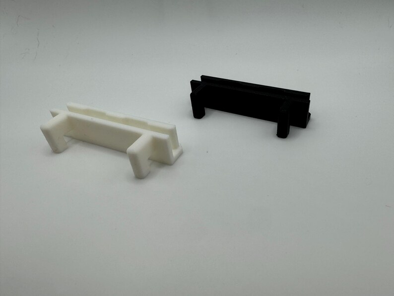 Pode incluir: Duas pe&ccedil;as de pl&aacute;stico, uma branca e outra preta, com um design semelhante. Ambas as pe&ccedil;as t&ecirc;m uma forma retangular com uma sec&ccedil;&atilde;o elevada no meio e duas pequenas pernas de cada lado.
