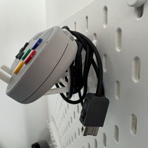 Puede incluir: Un mando blanco de Nintendo 64 con un cable negro colgando de una pared de tablero de clavijas blanco.