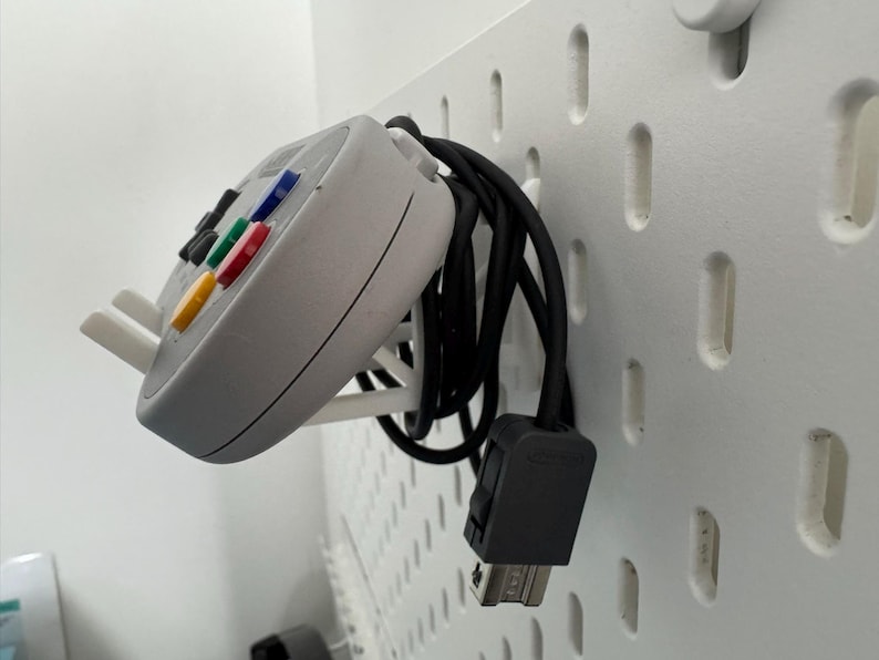 Puede incluir: Un mando blanco de Nintendo 64 con un cable negro colgando de una pared de tablero de clavijas blanco.