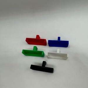 May include: Five plastic drawer pulls in different colours: red, blue, green, white and black. Each pull has a rectangular shape with a small handle on top.