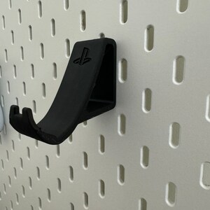 May include: A black plastic wall mount for a PlayStation controller. The mount has a PlayStation logo and a hook for hanging the controller.