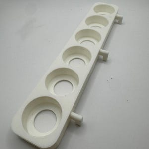 May include: A white, rectangular foam product with six circular indentations. The product has a series of circular holes and two small rectangular protrusions on the sides. The object is designed to hold multiple small, round objects.