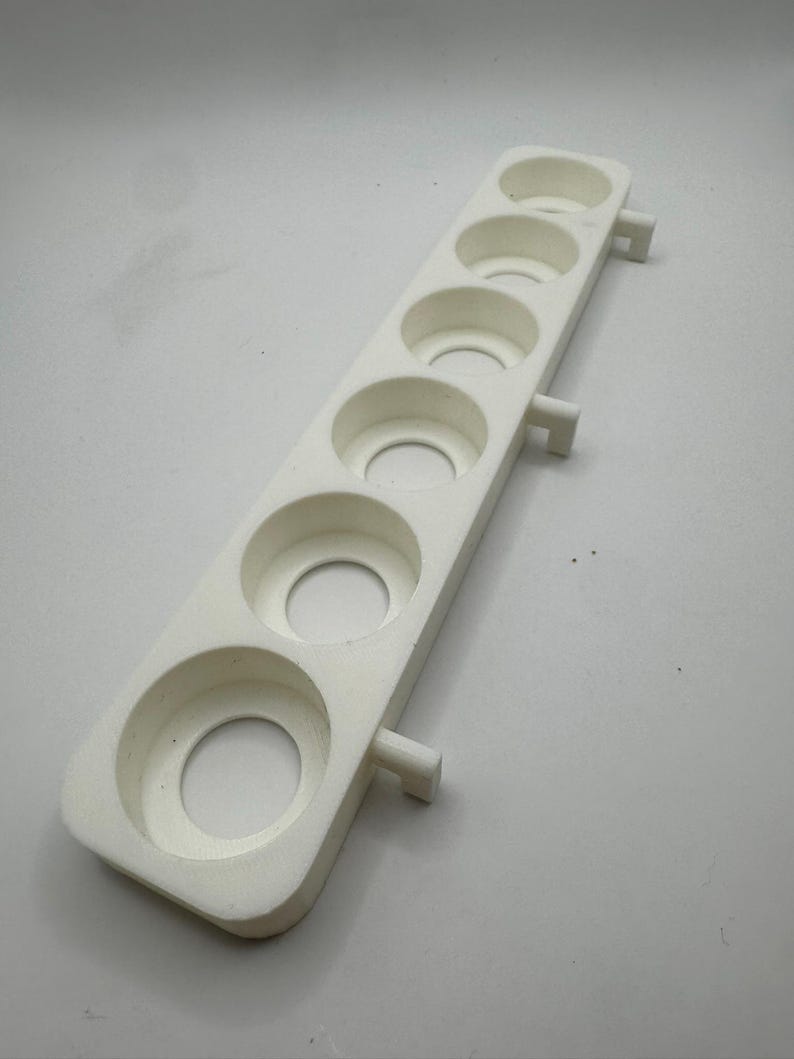 May include: A white, rectangular foam product with six circular indentations. The product has a series of circular holes and two small rectangular protrusions on the sides. The object is designed to hold multiple small, round objects.