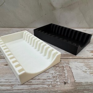 Op de afbeelding: Twee 3D-geprinte organizers, &eacute;&eacute;n wit en &eacute;&eacute;n zwart, staan naast elkaar. De witte organizer heeft een rechthoekige basis met een gebogen rand en verticale verdelers. De zwarte organizer heeft een vergelijkbaar ontwerp.