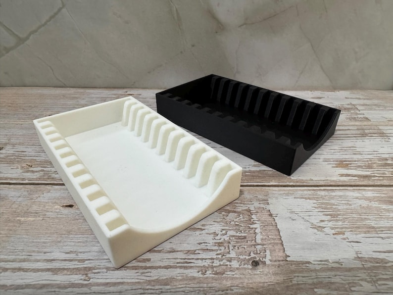 Op de afbeelding: Twee 3D-geprinte organizers, &eacute;&eacute;n wit en &eacute;&eacute;n zwart, staan naast elkaar. De witte organizer heeft een rechthoekige basis met een gebogen rand en verticale verdelers. De zwarte organizer heeft een vergelijkbaar ontwerp.