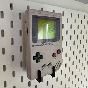 May include: A vintage Nintendo Game Boy handheld console in light gray, mounted on a white pegboard. The Game Boy features a black directional pad, red buttons, and the Nintendo logo. The screen reflects a green hue.