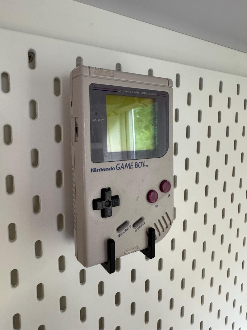 May include: A vintage Nintendo Game Boy handheld console in light gray, mounted on a white pegboard. The Game Boy features a black directional pad, red buttons, and the Nintendo logo. The screen reflects a green hue.