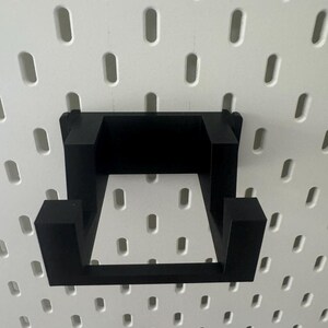 K&ouml;nnte beinhalten: Schwarzes, quadratisches Wandregal aus Kunststoff. Das Regal ist an einer wei&szlig;en Lochwand befestigt.