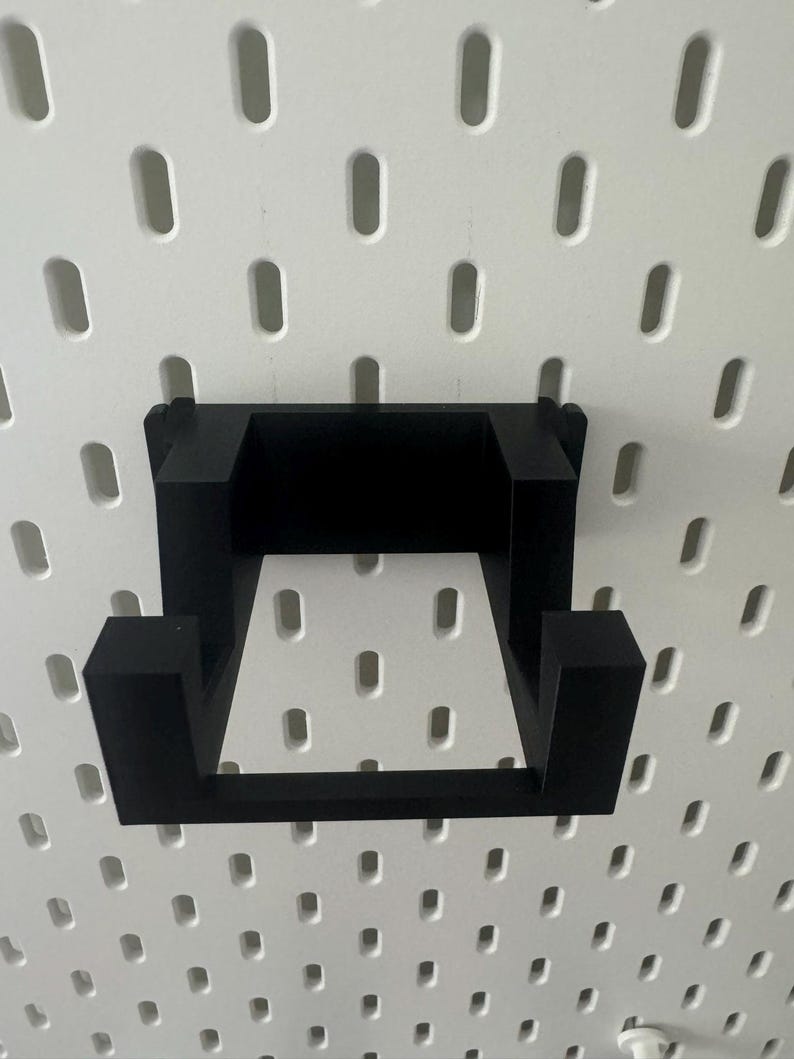 K&ouml;nnte beinhalten: Schwarzes, quadratisches Wandregal aus Kunststoff. Das Regal ist an einer wei&szlig;en Lochwand befestigt.