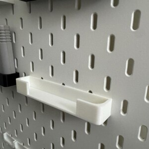 May include: A white pegboard with a white shelf and a video game cartridge. The shelf is rectangular and designed to hold small items. The pegboard has evenly spaced holes for attaching accessories.
