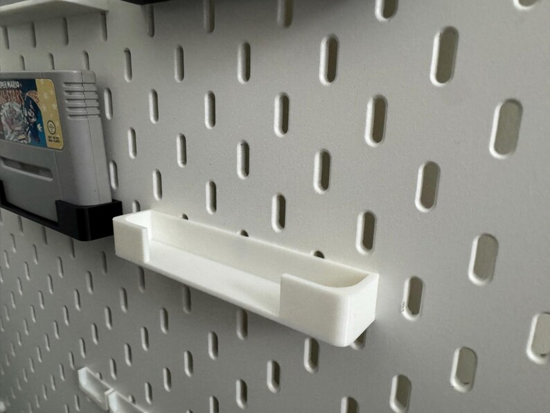 May include: A white pegboard with a white shelf and a video game cartridge. The shelf is rectangular and designed to hold small items. The pegboard has evenly spaced holes for attaching accessories.