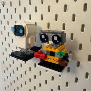 May include: A white robot with blue eyes and a yellow robot with black eyes and the word "WALL-E" on it. Both robots are made of Lego bricks and are displayed on a pegboard.