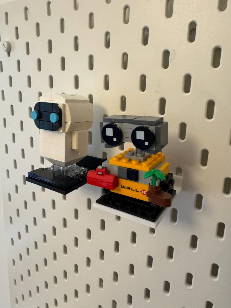 May include: A white robot with blue eyes and a yellow robot with black eyes and the word "WALL-E" on it. Both robots are made of Lego bricks and are displayed on a pegboard.