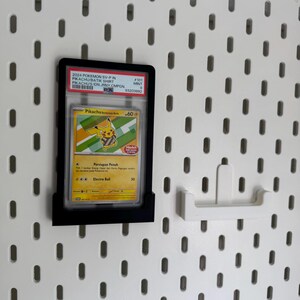 May include: A framed Pok&eacute;mon trading card featuring Pikachu, displayed on a white pegboard. The card is in a black holder and includes text such as "2024 Pokemon SV-P" and "Pikachu Batik Shirt". A white pegboard hook is also visible.